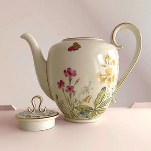 Może przedstawiać: Kremowa porcelanowa czajniczek z pokrywką, ozdobiona ilustracjami kwiatów i motyli. Czajniczek posiada zakrzywioną wylewkę, duży uchwyt i złote wykończenia. Pokrywka ma złoty uchwyt. Projekt zawiera różowe, żółte i niebieskie kwiaty.
