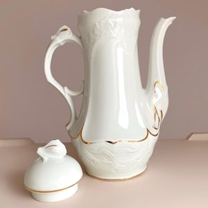 May include: A white porcelain coffee pot with a lid, featuring a curved handle and a long spout. The pot is adorned with gold trim and embossed floral patterns. The lid is placed next to the pot. The background is a soft pink.