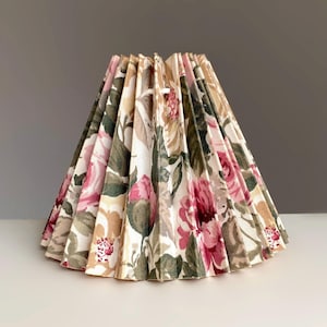 May include: A pleated lampshade with a floral design. The fabric displays pink roses, green leaves, and beige and brown tones on a white backdrop. The lampshade is cone-shaped.