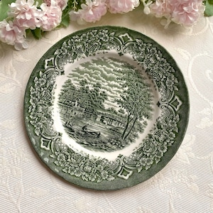 May include: A round ceramic plate featuring a dark green transferware pattern. The center depicts a landscape with a river, a small boat, and trees. The wide border is decorated with intricate floral motifs and a lattice design in dark green.
