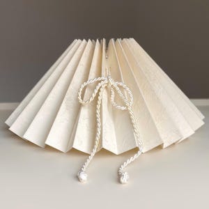 May include: A cream-colored pleated lampshade with a conical shape and a subtle textured pattern. The shade features a decorative white twisted rope cord tied into a bow at the front, with two long ends hanging down, each finished with a small knot.