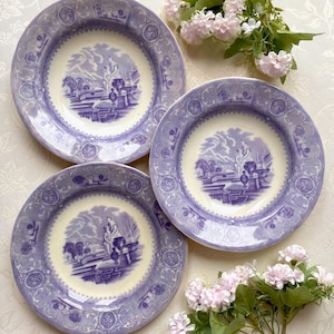May include: Three antique, round plates with a lavender and cream color scheme. Each plate features a central landscape scene in lavender and a decorative border. The plates are arranged with pink flowers.