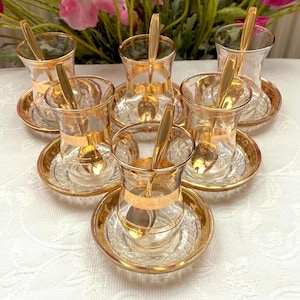 Puede incluir: Un juego de seis tazas de té de vidrio transparente con bordes dorados y platillos a juego. Cada taza tiene una cuchara dorada en su interior. Las tazas y los platillos están dispuestos sobre una superficie blanca.