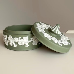 May include: A sage green round ceramic lidded jar featuring white raised relief detailing of grapevines and leaves encircling the base and the lid. The lid includes a pointed, textured finial with white leaf patterns on a sage green background.