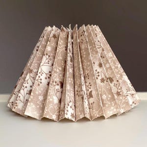 May include: A pleated lampshade with a floral pattern in beige and brown. The shade has a conical shape and is made of fabric. The floral design includes small flowers and leaves, creating a delicate and natural aesthetic.