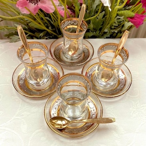 Puede incluir: Juego de cuatro tazas de té de vidrio transparente con bordes dorados y platillos a juego. Cada taza tiene una cuchara dorada en su interior. Los platillos y las tazas tienen un diseño decorativo dorado y plateado.