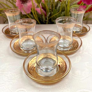 Puede incluir: Un juego de cinco tazas de té de cristal transparente con sus platos a juego, cada una con una silueta curva y bordes decorativos en tono dorado. Los bordes de las tazas y los platos están adornados con intrincados patrones dorados y filigranas.