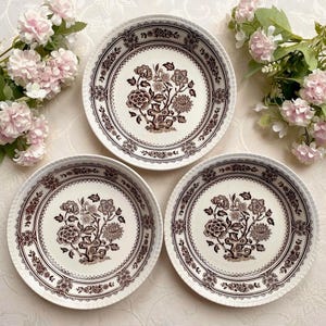 May include: Three round ceramic plates featuring a dark brown floral pattern on a cream background. Each plate displays a central botanical illustration of a flowering tree surrounded by a decorative border with intricate floral and geometric motifs.