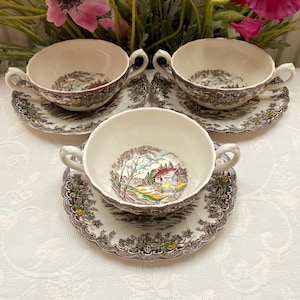 May include: Three cream-coloured ceramic soup bowls with two handles each, paired with matching saucers. The bowls and saucers feature a brown transferware landscape scene depicting a house, trees, and fields, with scalloped edges.