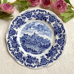 May include: A white ceramic plate featuring a blue transferware pattern. The centre depicts a castle landscape with a river and clouds. The rim is decorated with a detailed floral and scrollwork border. The plate is surrounded by pink artificial roses.