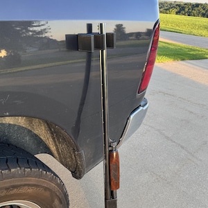 May include: A black shotgun is mounted vertically on the side of a gray truck. The shotgun has a brown wooden stock and forend. A black mounting bracket is attached to the truck's body.