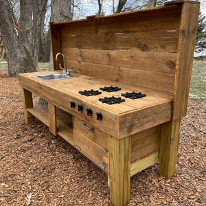 May include: A wooden play kitchen with a sink, stovetop, and storage drawers. The kitchen is made from reclaimed wood and has a rustic look. It is perfect for outdoor play.