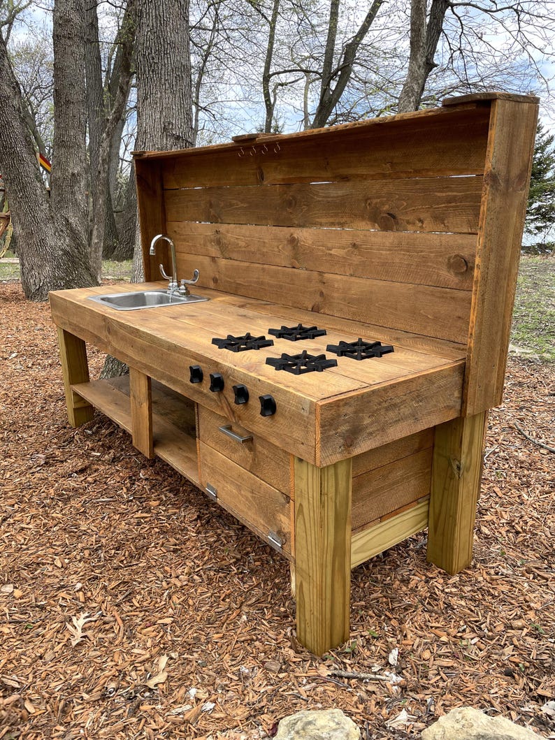 May include: A wooden play kitchen with a sink, stovetop, and storage drawers. The kitchen is made from reclaimed wood and has a rustic look. It is perfect for outdoor play.