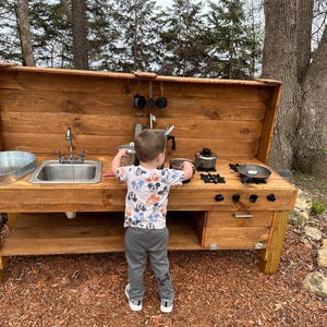 May include: A wooden outdoor play kitchen with a sink, stovetop, and storage shelf. A young child is playing with the kitchen, pretending to cook.