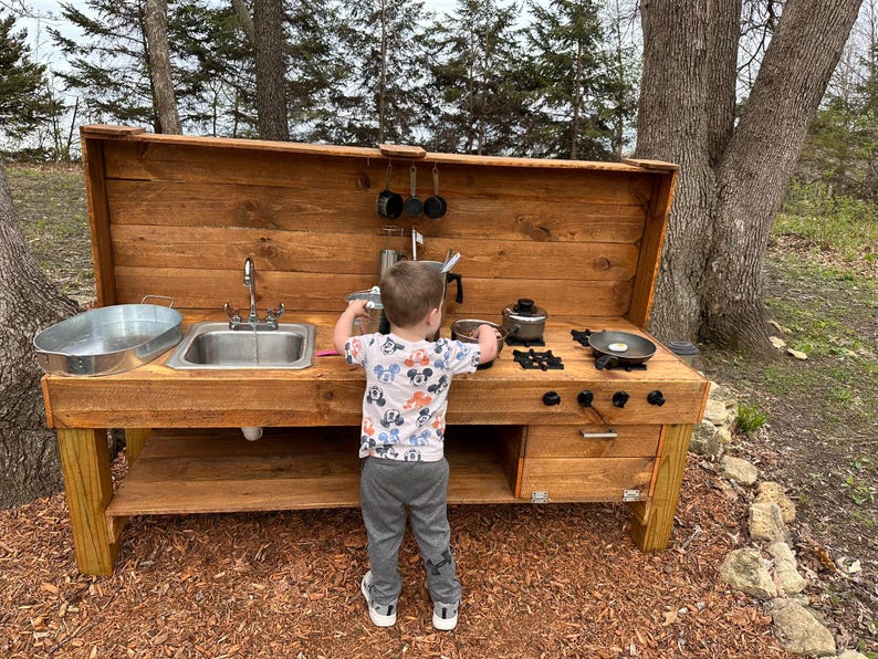 May include: A wooden outdoor play kitchen with a sink, stovetop, and storage shelf. A young child is playing with the kitchen, pretending to cook.