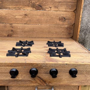 May include: A rustic wooden outdoor kitchen with four black cast iron burner grates and three black knobs. The kitchen has a drawer with a silver handle.