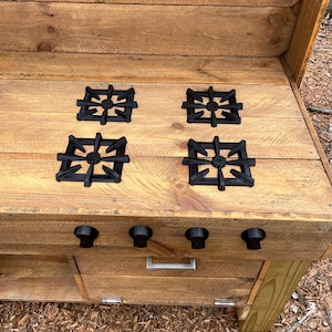 May include: A wooden play kitchen stove with four black burner covers and three black knobs. The stove has a wooden drawer and a wooden shelf below the stovetop.