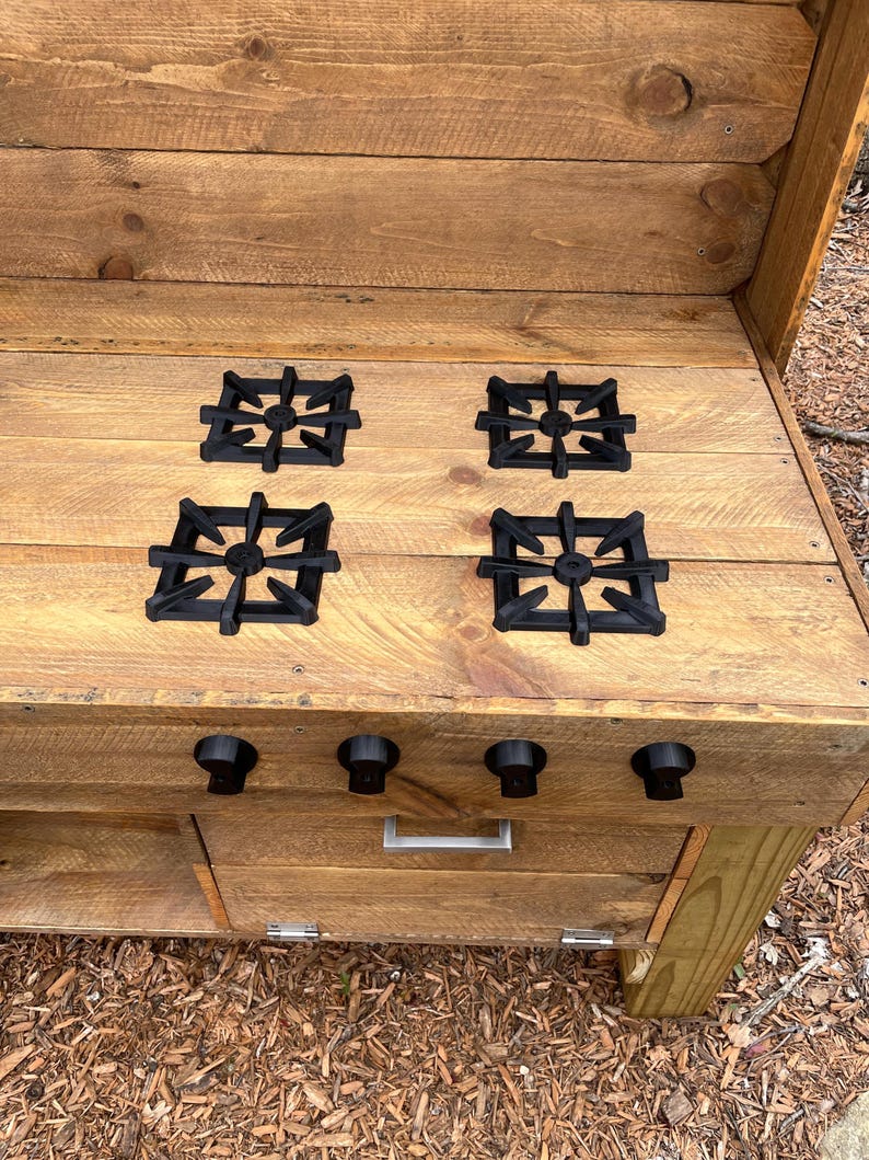 May include: A wooden play kitchen stove with four black burner covers and three black knobs. The stove has a wooden drawer and a wooden shelf below the stovetop.