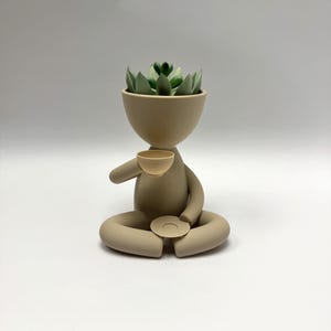 May include: A beige ceramic planter shaped like a person sitting in a meditative pose. The planter has a small cup in one hand and a saucer in the other. A green succulent plant is growing in the planter's head.