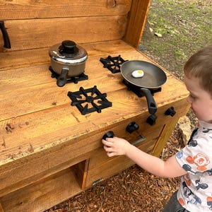 May include: A wooden play kitchen with a black stovetop and a child's hand reaching for a knob. The stovetop has two burners with black metal grates and a silver pot and a black frying pan with a white egg on it.