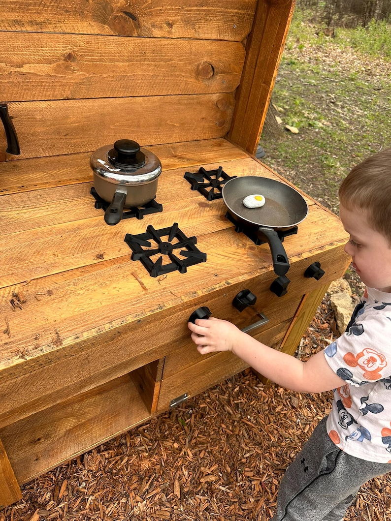 May include: A wooden play kitchen with a black stovetop and a child's hand reaching for a knob. The stovetop has two burners with black metal grates and a silver pot and a black frying pan with a white egg on it.
