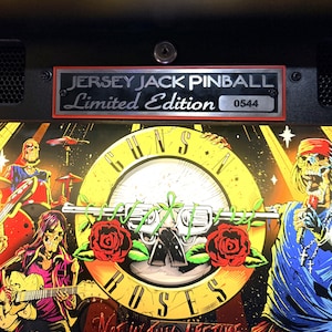 May include: A pinball machine featuring the Guns N' Roses band logo with a central revolver design, roses, and the text "Not In This Lifetime." The machine has a "Jersey Jack Pinball Limited Edition" plaque with the number 0544.