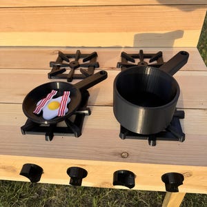 May include: A wooden play kitchen with a black stovetop and burners. A black frying pan with a toy egg and bacon sits on one burner. A black pot sits on another burner. The kitchen is made of light-colored wood.
