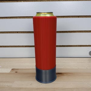 May include: A red can cooler with vertical ridges, resembling a shotgun shell, holds a yellow can with "COLLECTOR'S EDITION" text. The base of the cooler is dark gray. The cooler sits on a wooden surface.