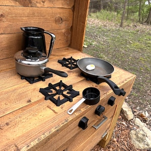 May include: A wooden play kitchen with a black stovetop, a black pitcher, a silver pot, a black frying pan, a white egg, a black measuring cup, and a silver spoon. The kitchen has a wooden drawer and a wooden door.