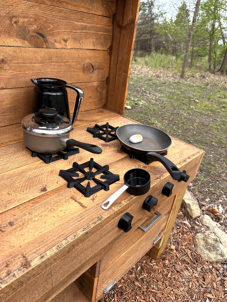 May include: A wooden play kitchen with a black stovetop, a black pitcher, a silver pot, a black frying pan, a white egg, a black measuring cup, and a silver spoon. The kitchen has a wooden drawer and a wooden door.