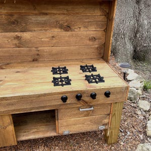 May include: A wooden outdoor kitchen with a built-in stovetop. The stovetop has four black cast iron burner grates and a drawer below. The kitchen is made of rustic wood planks and has a backsplash.
