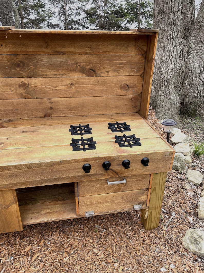 May include: A wooden outdoor kitchen with a built-in stovetop. The stovetop has four black cast iron burner grates and a drawer below. The kitchen is made of rustic wood planks and has a backsplash.