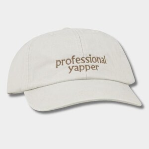 May include: An off-white baseball cap featuring the text professional yapper embroidered in brown thread across the front panel. The cap has a curved brim, a structured crown, and a fabric strap with a metal buckle closure at the back.
