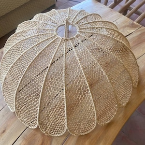 Könnte beinhalten: Ein gewebter, beigefarbener Lampenschirm mit einem abgerundeten, gewellten Design. Der Lampenschirm besteht aus einem netzartigen Material, das ein zartes, luftiges Aussehen erzeugt. Der Lampenschirm ist für die Platzierung auf einem Tisch oder einer anderen Oberfläche konzipiert.