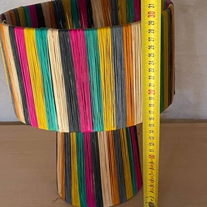 May include: A colorful lamp with a cylindrical top and a conical base. The lamp is made of woven strips of colorful material, including yellow, orange, green, pink, black, and white.