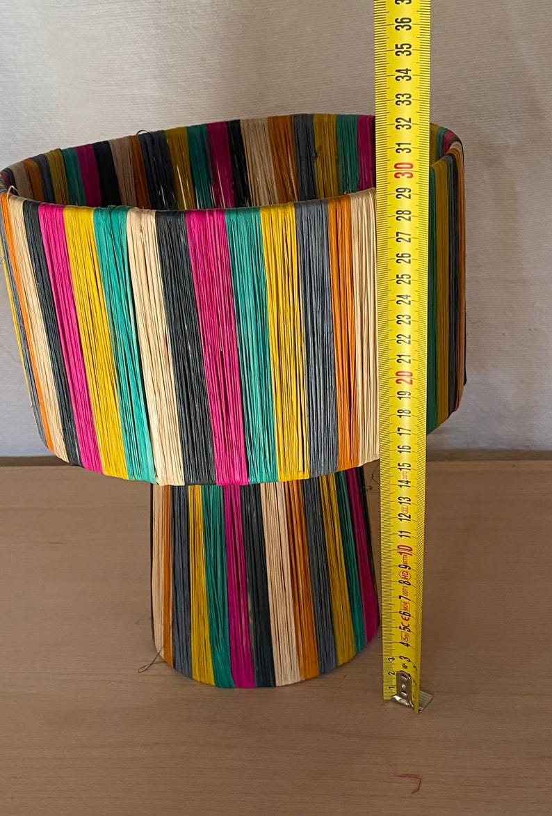 May include: A colorful lamp with a cylindrical top and a conical base. The lamp is made of woven strips of colorful material, including yellow, orange, green, pink, black, and white.