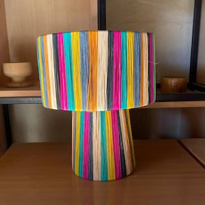 May include: A colorful lamp with a striped shade and base made of woven natural fibers. The lamp is on a wooden table with a woven wicker bottom.