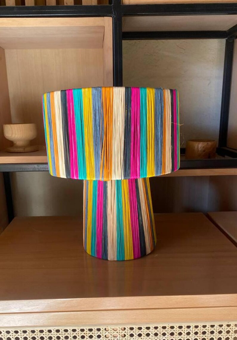 May include: A colorful lamp with a striped shade and base made of woven natural fibers. The lamp is on a wooden table with a woven wicker bottom.