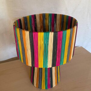 May include: A colorful woven lamp base with a wide, cylindrical top and a smaller, round base. The base is made of natural fibers and is decorated with stripes of various colors, including black, white, pink, yellow, green, and orange.