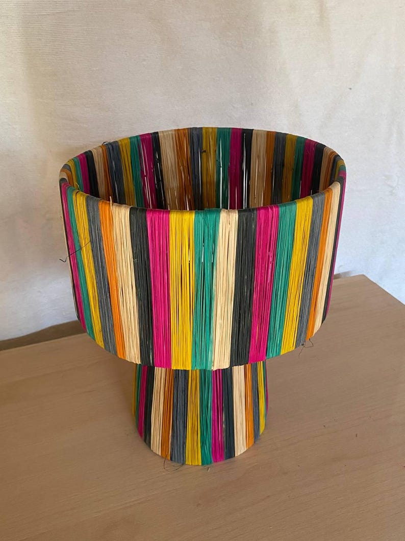 May include: A colorful woven lamp base with a wide, cylindrical top and a smaller, round base. The base is made of natural fibers and is decorated with stripes of various colors, including black, white, pink, yellow, green, and orange.