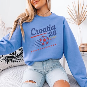 A woman wearing a blue sweatshirt with a graphic design of a soccer ball and the text "Croatia '26" is sitting on a bed.