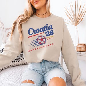 A woman wearing a beige sweatshirt with a graphic of a soccer ball and the text "Croatia 26" is sitting on a bed.