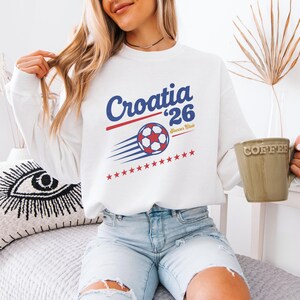 A woman wearing a white sweatshirt with a graphic of a soccer ball and the text "Croatia '26" is sitting on a bed, holding a coffee mug.