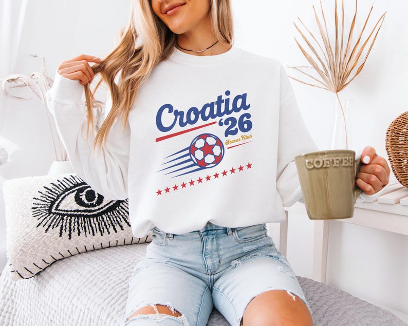 A woman wearing a white sweatshirt with a graphic of a soccer ball and the text "Croatia '26" is sitting on a bed, holding a coffee mug.