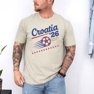 A man wearing a beige t-shirt with a graphic design of a soccer ball and stars, standing against a white wall.