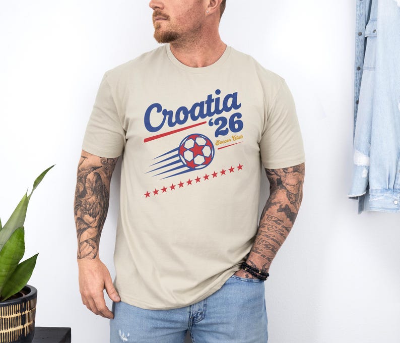 A man wearing a beige t-shirt with a graphic design of a soccer ball and stars, standing against a white wall.