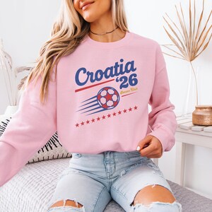 A woman wearing a pink sweatshirt with a graphic of a soccer ball and the text "Croatia '26" is sitting on a bed.