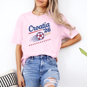 A woman wearing a pink t-shirt with a graphic of a soccer ball and the text "Croatia 26" is standing next to a potted plant.