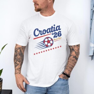A man wearing a white t-shirt with a graphic design of a soccer ball and stars, standing in front of a plant and a denim jacket.