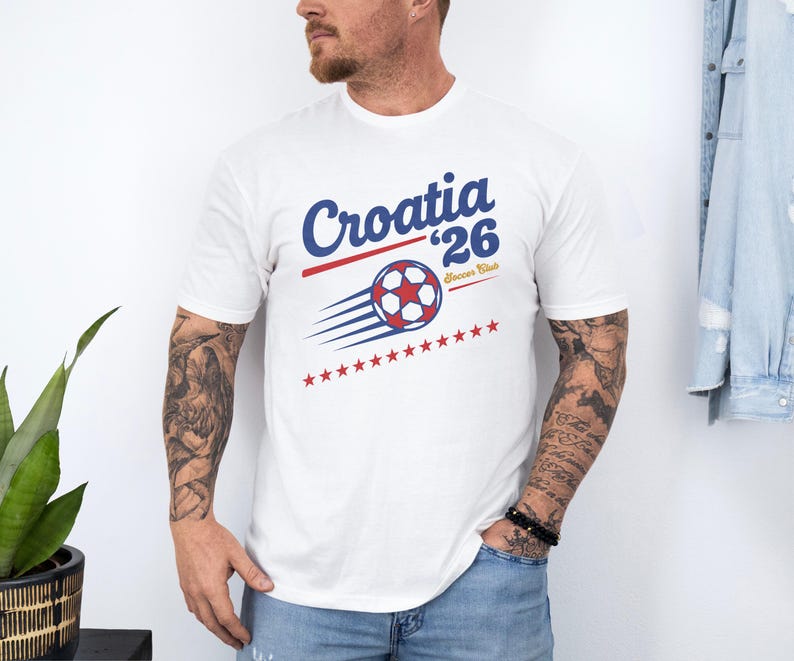 A man wearing a white t-shirt with a graphic design of a soccer ball and stars, standing in front of a plant and a denim jacket.
